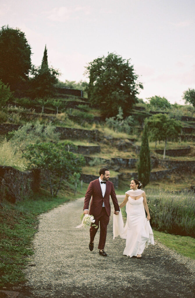 Bride and groom walk through lush green gardens, looking at each other and smiling, enjoying one of the best wedding venues in Sicily. 